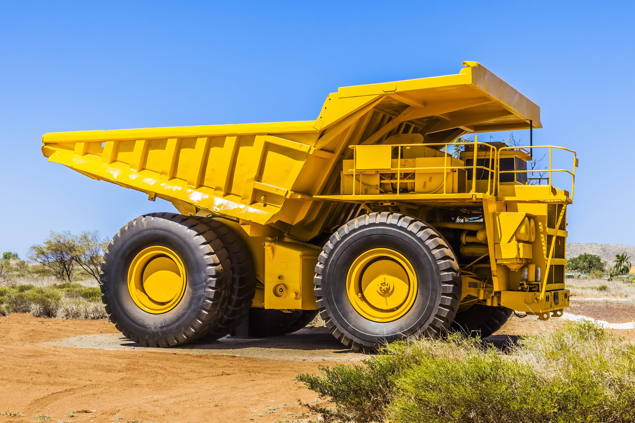 A large yellow dump truck is positioned on a sandy surface under a clear blue sky, showcasing its significant size and heavy-duty design for mining or construction.